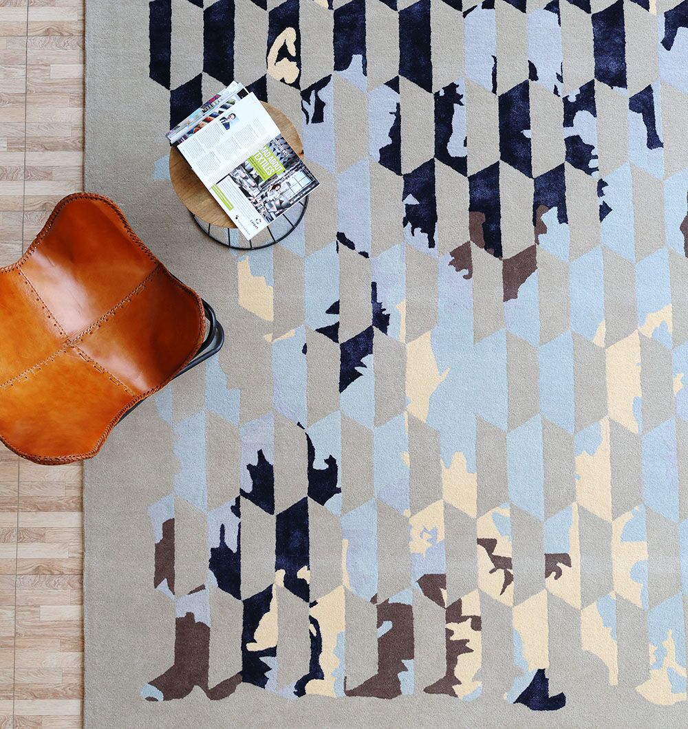A top-down view of the RAMSHA HOME Multi Color Handmade Decorative Area Rug - HTWV 166 (300X400 cm), featuring abstract blue, gray, beige, and brown patterns, styled with a brown leather chair and small round table with a magazine.