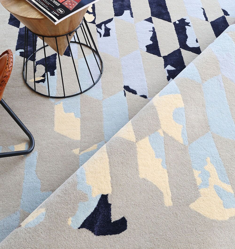 The RAMSHA HOME Multi Color Handmade Decorative Area Rug - HTWV 166 (300x400 cm) features abstract and diamond patterns in beige, light blue, navy, and cream, shown with an angled wooden side table and a partial metal chair.