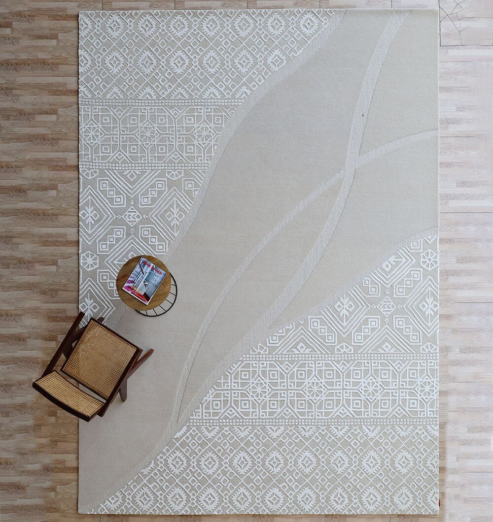 A top view of the RAMSHA HOME Boho Geometric Handmade Decorative Area Rug - HTWV 167 (300x400 cm) in beige with wavy lines on a light wooden floor; a woven chair and round side table with magazines appear in one corner.