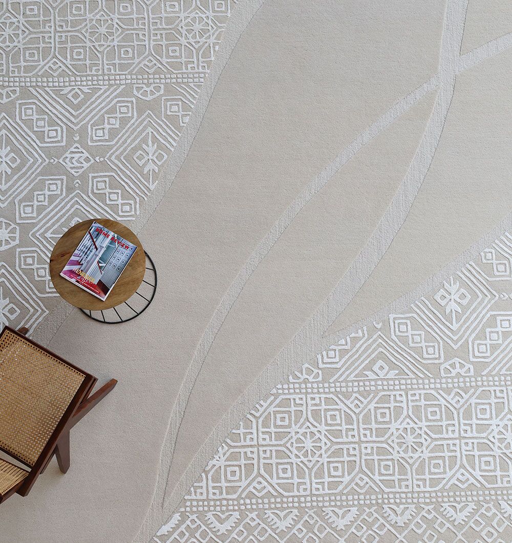 A small round wooden table with magazines and a woven chair rest on the RAMSHA HOME Boho Geometric Handmade Decorative Area Rug – HTWV 167 (300x400 cm), featuring intricate white geometric patterns and curved lines.