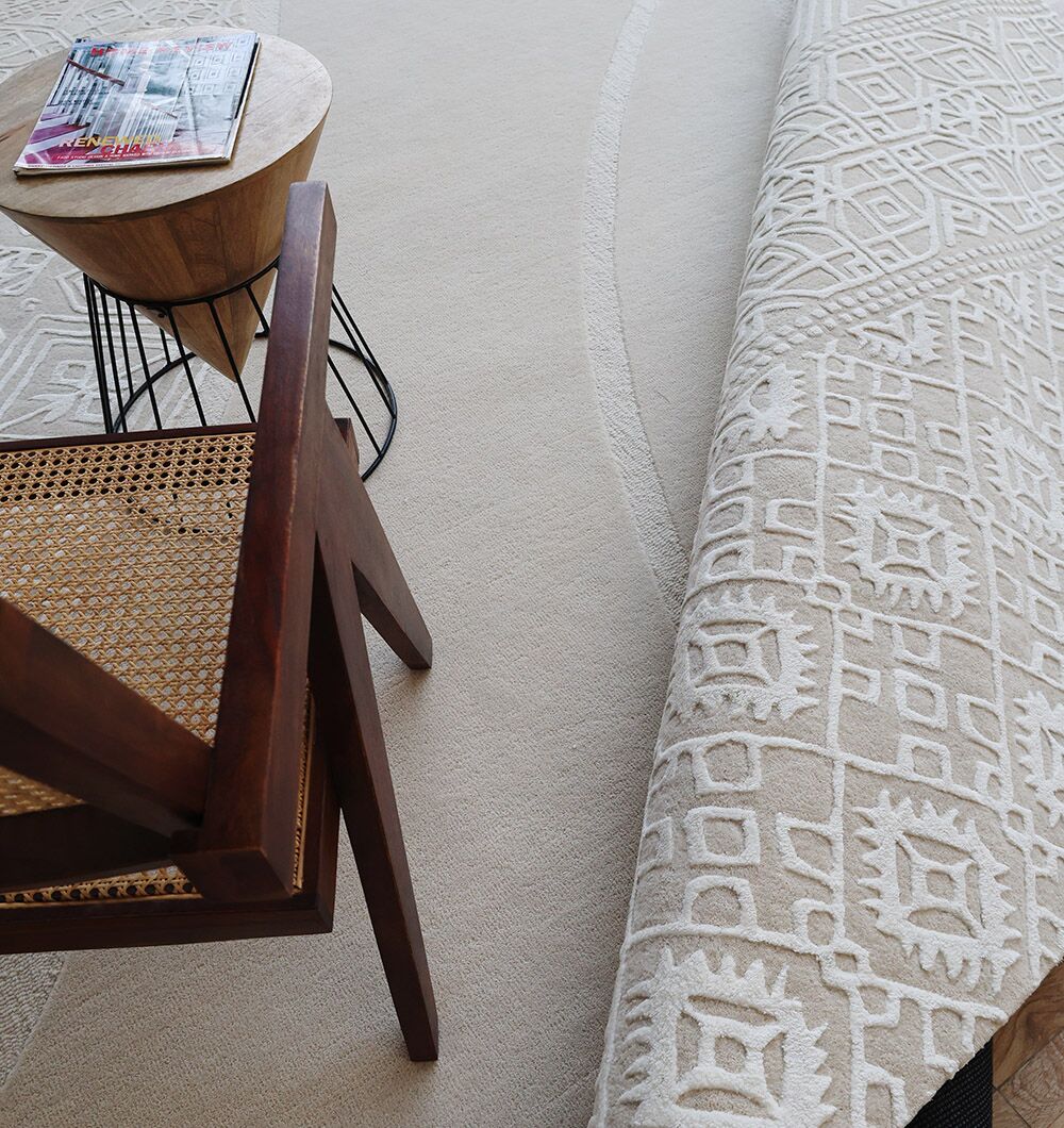 A wooden chair with a woven seat sits beside a round side table with magazines. A RAMSHA HOME Boho Geometric Handmade Decorative Area Rug – HTWV 167 (300x400 cm) in cream is partially rolled, showing the plain beige carpet beneath.
