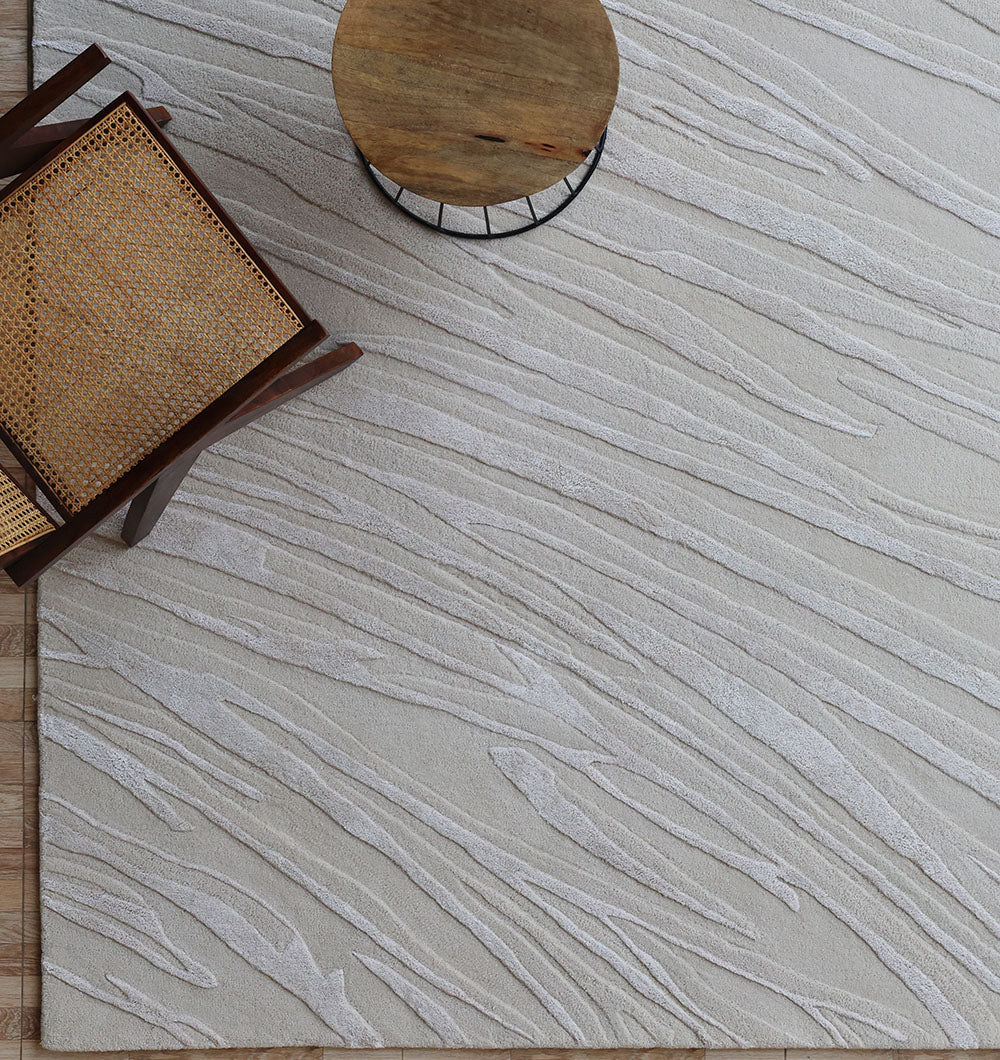 The Decorative Area Handmade Rug - HTWV 168 by RAMSHA HOME, featuring a light tone and subtle wavy texture, is displayed beneath a wooden chair with a woven cane seat and a round wooden side table.