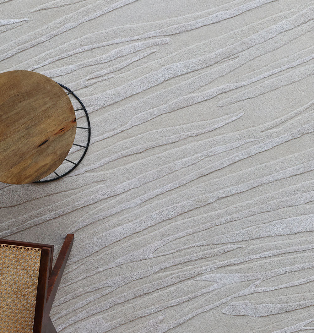 The Decorative Area Handmade Rug - HTWV 168 by RAMSHA HOME features a neutral, wavy textured pattern. A round wooden table and a woven-seat chair appear in the bottom left corner.