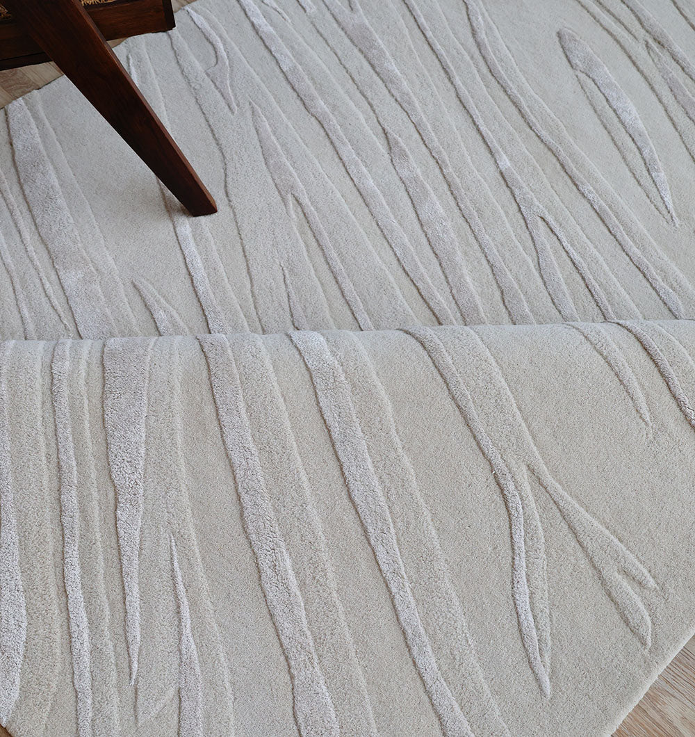 A close-up of the RAMSHA HOME Decorative Area Handmade Rug - HTWV 168 shows its cream color, hand-tufted wool, and raised wavy linear patterns, partially folded. A wooden furniture leg appears in the upper left corner.