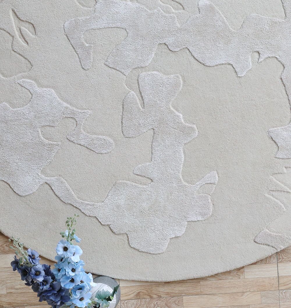 A close-up of the RAMSHA HOME Round Home Decor Handmade Rug - HTWV 170 (200 cm), featuring abstract raised patterns, displayed on a wooden floor. A small bouquet of blue and white flowers appears in the lower left corner.