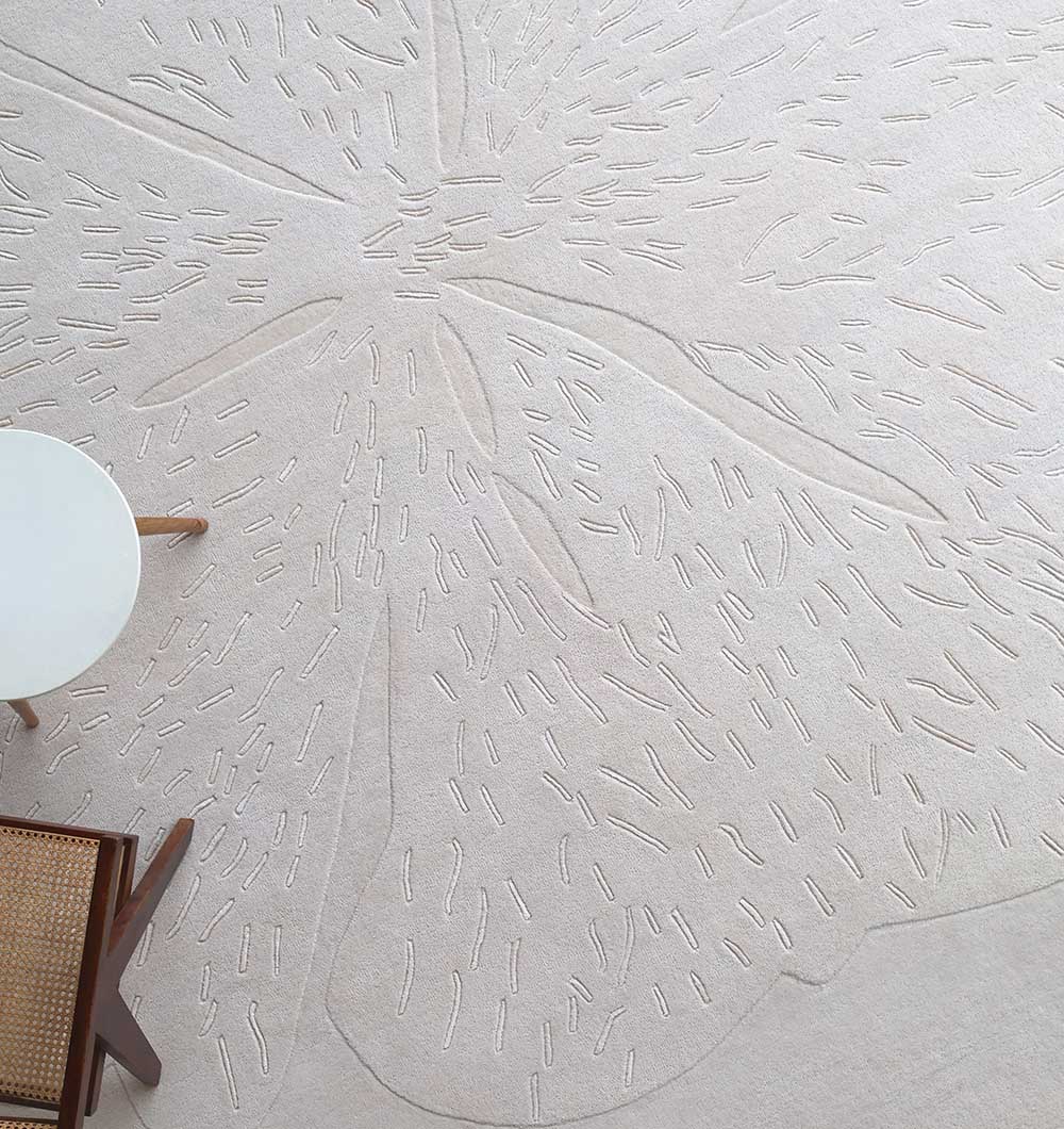 The RAMSHA HOME Embossed Floral Solid Light Grey Rug – HTWV 171 features a textured hand-tufted design with subtle patterns, placed beside a round white table and a wooden chair with a woven rattan seat.