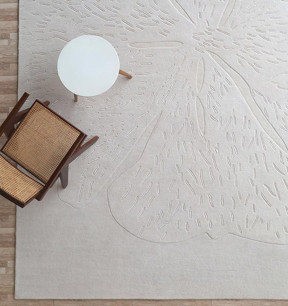 A top-down view of the RAMSHA HOME Embossed Floral Solid Light Grey Rug – HTWV 171 with abstract raised patterns, a round white side table, and a wooden chair with woven cane seat and back on a light wooden floor.