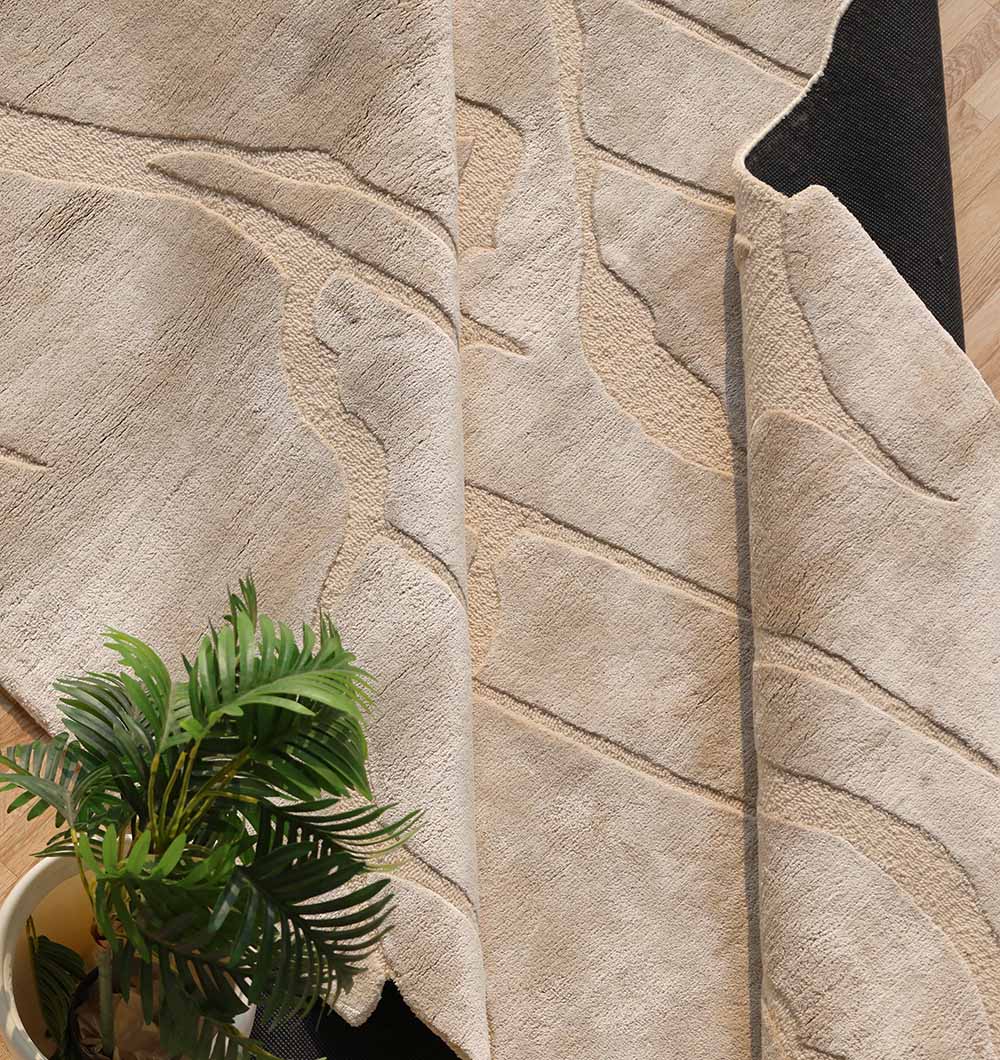 A close-up of the RAMSHA HOME Modern Viscose & Wool Handmade Carpet 200x300 – HTWV-172 shows its abstract beige design with a black underside, partially folded. A potted green plant rests on the light wood floor in the lower left corner.
