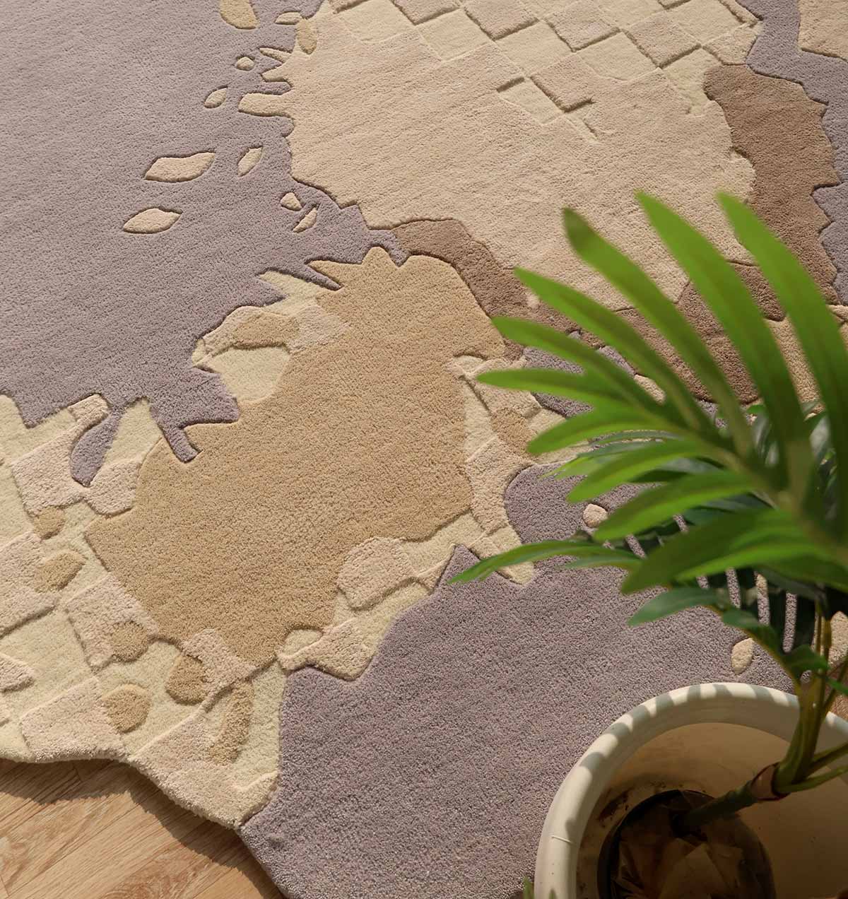 A potted green plant with slender leaves sits on a floor covered by the Abstract Shaped Handmade Area Rug-HTWV 174 (200x300 cm) from RAMSHA HOME, featuring textured, multi-colored beige and lavender abstract patterns.