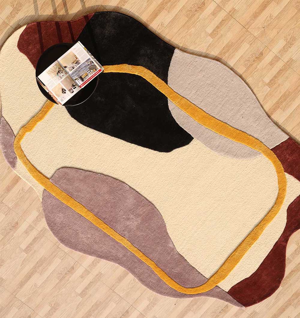 The Handmade irregular Multi Color Carpet HTWV-176 by RAMSHA HOME, featuring abstract wavy shapes in black, beige, yellow, brown, and gray tones, is displayed on a wood floor next to a small round table with books and magazines.