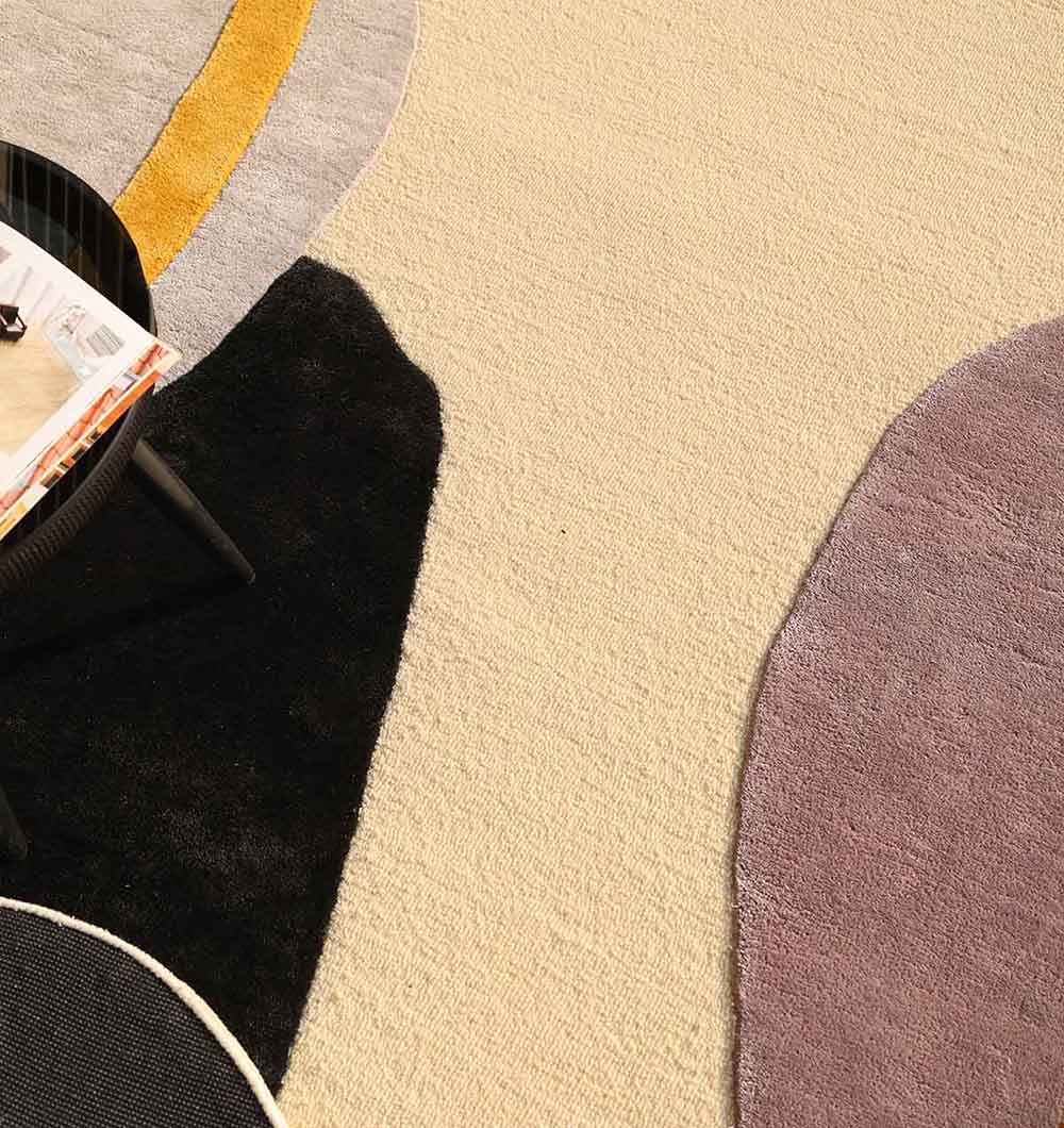 The RAMSHA HOME Handmade Irregular Multi Color Carpet HTWV-176, a hand-tufted wool rug with abstract black, gray, purple, yellow, and cream shapes, is shown beneath a round table with magazines in the upper left corner.