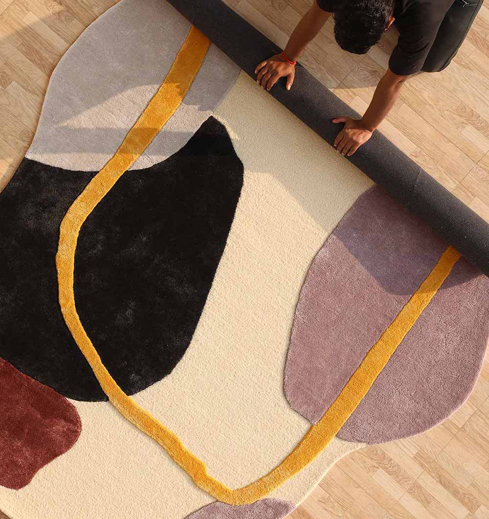 A person unrolls the RAMSHA HOME Handmade irregular Multi Color Carpet HTWV-176, featuring abstract black, maroon, mauve, cream, and yellow shapes on a light wood floor.