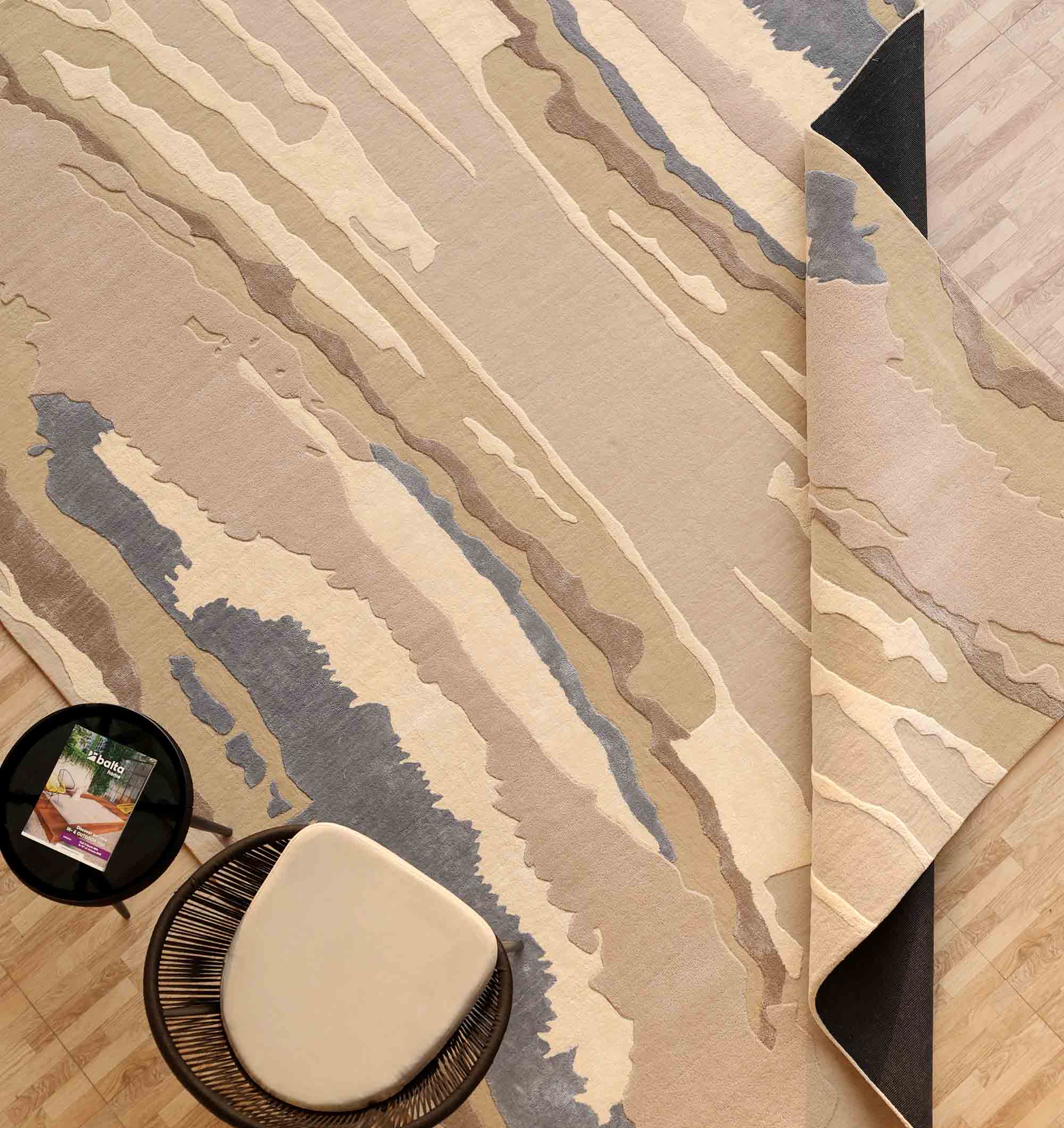 The RAMSHA HOME Classic Rectangle Handmade Area Rug- HTWV 177 (300x400 cm) in neutral beige, tan, and gray is partially folded on a light wood floor beside a round side table with a book and a woven chair with a light cushion.