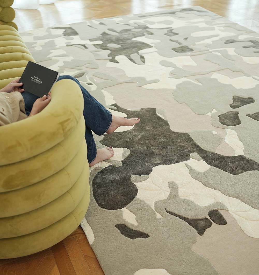 Barefoot, a person sits on a light green sofa with a book, their feet resting on the RAMSHA HOME Abstract Wool & Viscose Handmade Carpet 300x400 (HTWV-180) in neutral abstract tones atop the wooden floor.