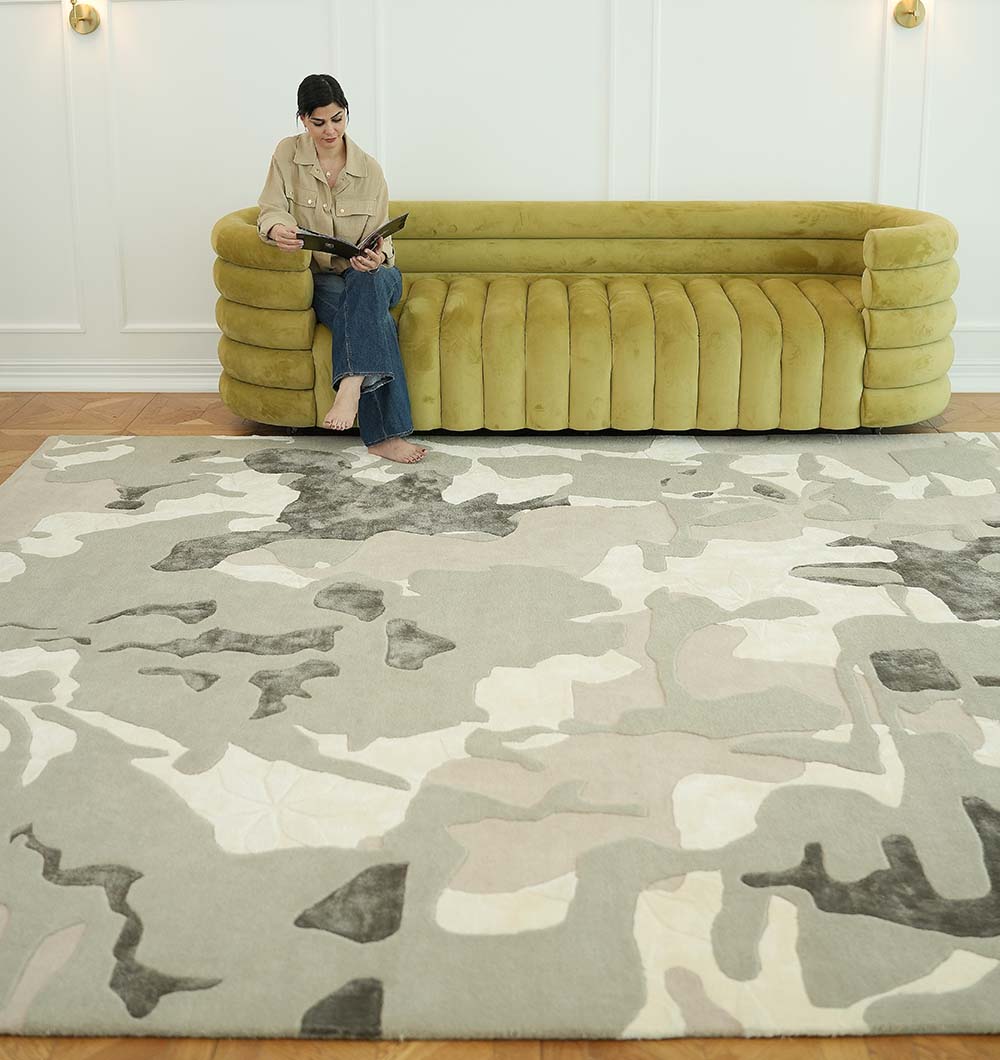 A woman reads on a modern olive-green sofa with bare feet resting on the RAMSHA HOME Abstract Wool & Viscose Handmade Carpet 300x400 – HTWV-180 in a bright room featuring white walls and wooden floors.