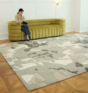 A woman sits barefoot on a green modern sofa reading, with the RAMSHA HOME Abstract Wool & Viscose Handmade Carpet 300x400 – HTWV-180 in neutral tones spread out before her on a parquet wood floor in a bright, minimal room.