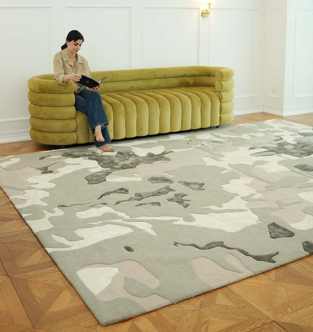 A woman sits barefoot on a green modern sofa reading, with the RAMSHA HOME Abstract Wool & Viscose Handmade Carpet 300x400 – HTWV-180 in neutral tones spread out before her on a parquet wood floor in a bright, minimal room.