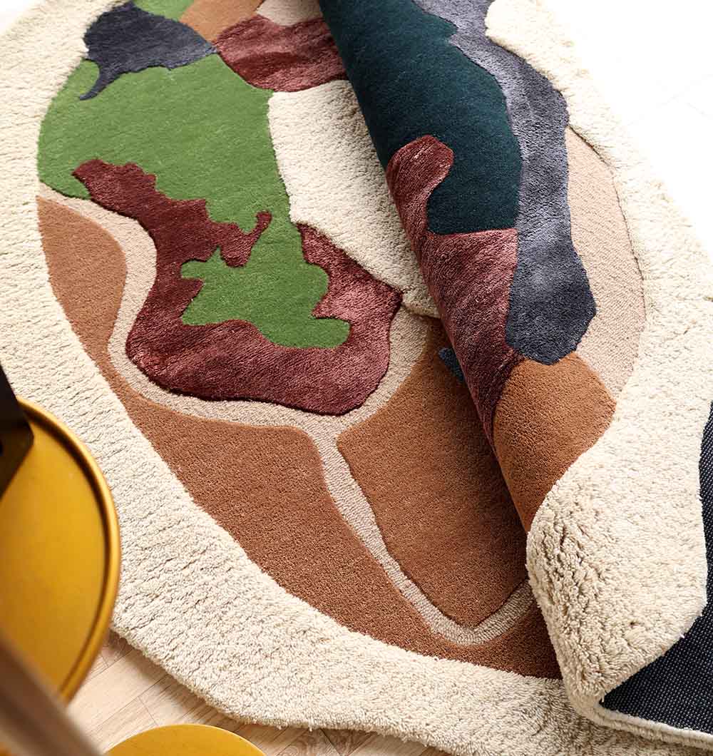 The Handmade Multicolor Abstract Shaped Rug HTWV 181 (200cm) by RAMSHA HOME, with green, brown, cream, and black earth-toned shapes, is partially rolled to reveal its textured surface; a yellow chair leg contrasts against this modern rug.