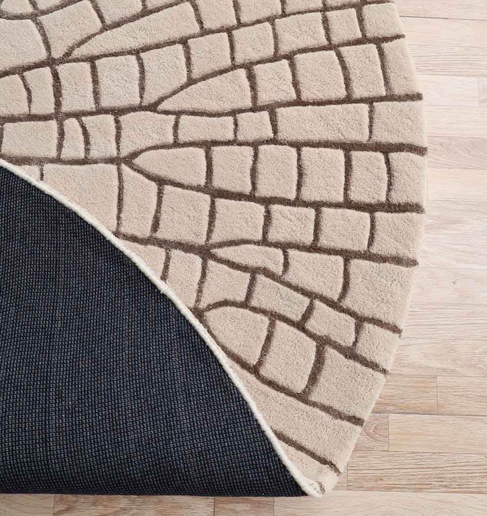 The RAMSHA HOME Beige & Brown Decorative Handmade Shaped Rug HTWV 182 (200 cm) features a modern geometric, stone-like pattern. Partially flipped, it reveals a dark textured underside while resting on a light wooden floor.