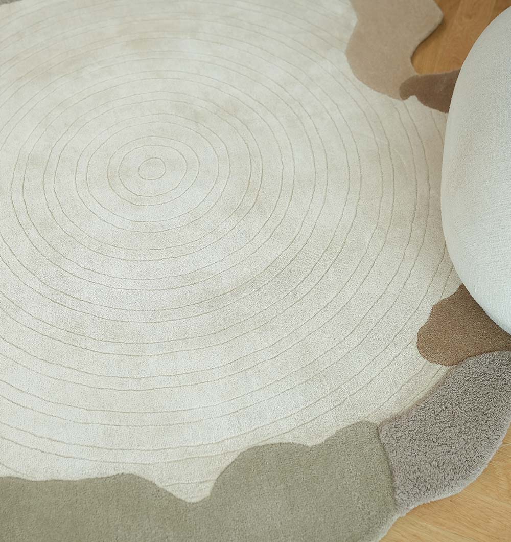 The RAMSHA HOME Decorative Handmade Modern Shaped Rug HTWV 185 (183 x 200 cm), featuring cream tones with concentric circles and uneven beige-brown edges like a tree trunk, lies on a wooden floor beside a curved pale cushion.
