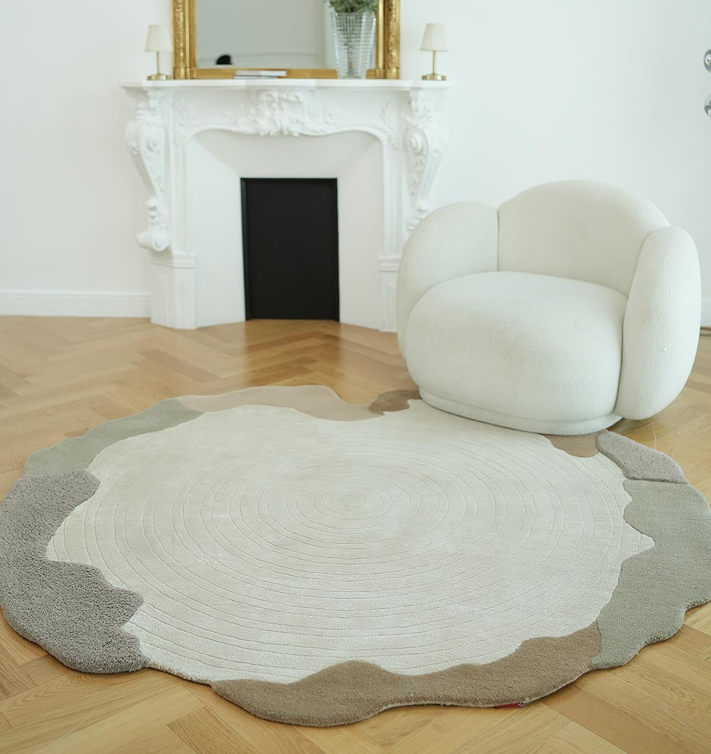 A modern living room featuring the RAMSHA HOME Decorative Handmade Modern Shaped Rug HTWV 185 (183 x 200 cm) in neutral tones, a plush white armchair, and an ornate white fireplace with a gold-framed mirror on the mantel.