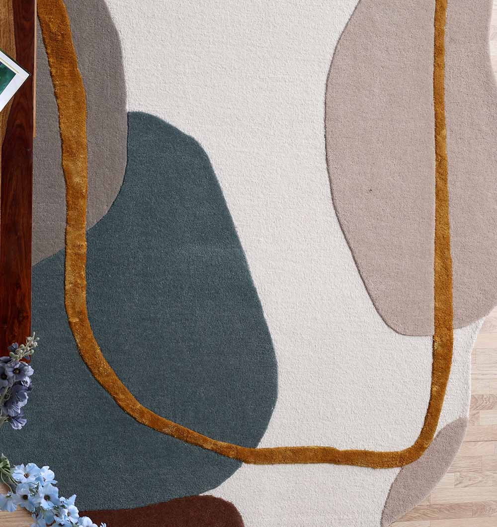 The RAMSHA HOME Handmade Multi Color Shaped Area Rug 200x300 CM HTWV 186 features abstract overlapping shapes in cream, dark green, beige, and brown with mustard yellow accents. A wooden table and a bouquet appear in the bottom left corner.
