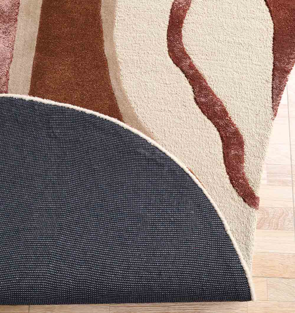 The Decorative Handmade Multi Color Shaped Area Rug – HTWV 187 by RAMSHA HOME features curved brown and beige patterns on a light wood floor. A lifted corner reveals a dark, textured rug pad, highlighting its premium hand tufted wool quality.