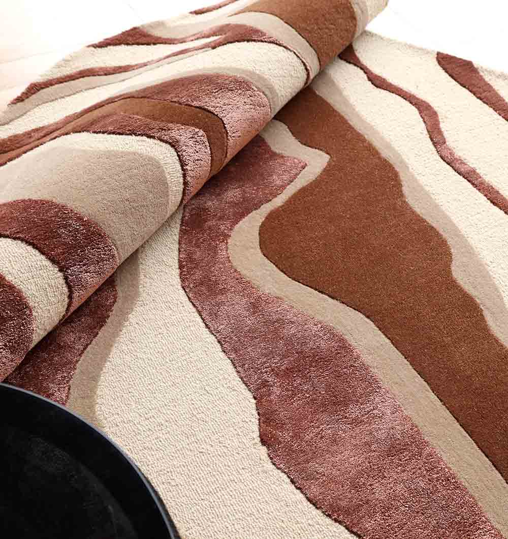 A close-up of the Decorative Handmade Multi Color Shaped Area Rug – HTWV 187 by RAMSHA HOME shows its wavy abstract brown, beige, and cream patterns. One edge is partially rolled up to reveal the rich hand-tufted wool texture.