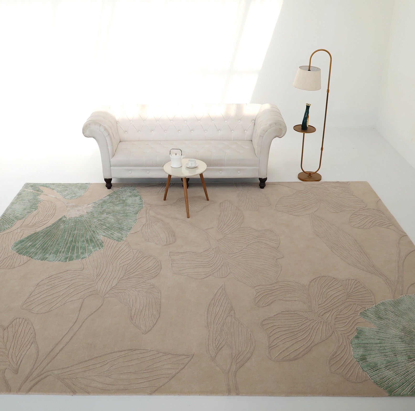 A minimalist living room with a white tufted sofa, modern floor lamp, two round side tables, and the RAMSHA HOME Floral Handmade Rectangle Rug HTWV-189, featuring subtle green leaf accents. Sunlight streams in from the left.