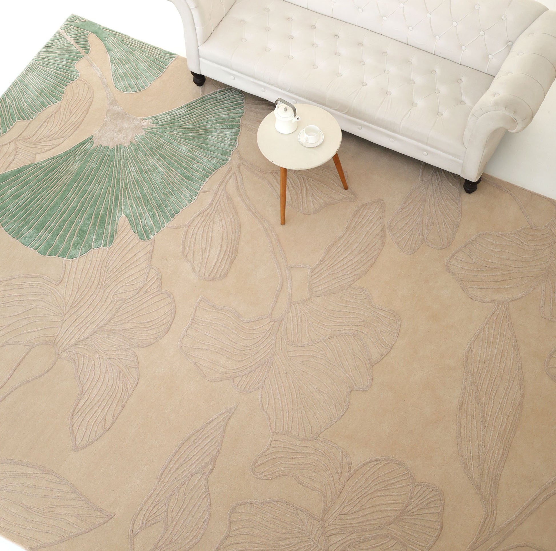 A cream sofa and small round table with a teapot and cup sit on the RAMSHA HOME Floral Handmade Rectangle Rug HTWV-189, featuring large textured flowers and a bold green leaf design.