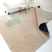 The RAMSHA HOME Floral Handmade Rectangle Rug HTWV-189, in beige with large pale green leaf designs, lies partially folded on a white floor. In the background are a white sofa, round side table with tea set, and floor lamp.