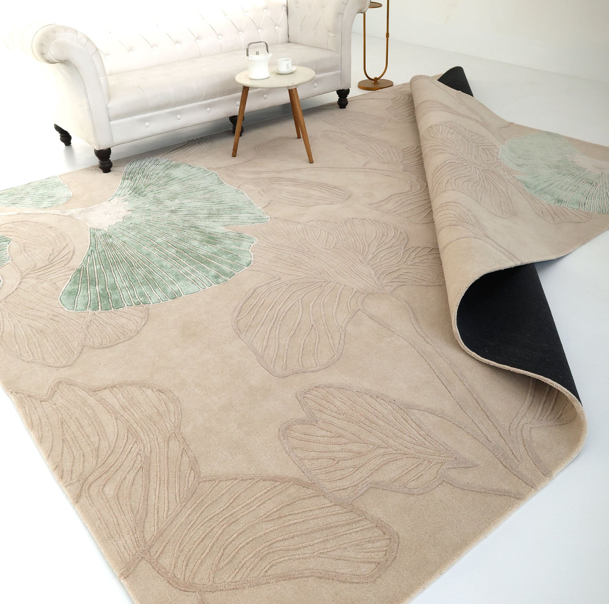 The RAMSHA HOME Floral Handmade Rectangle Rug HTWV-189, in beige with large pale green leaf designs, lies partially folded on a white floor. In the background are a white sofa, round side table with tea set, and floor lamp.