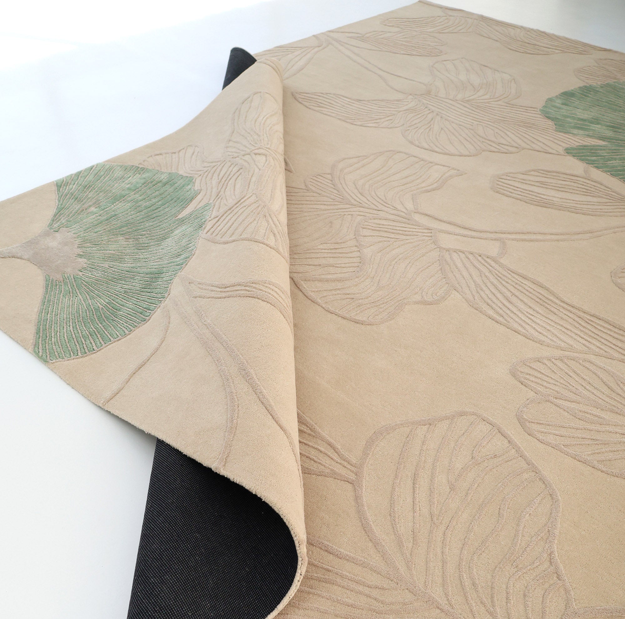 The RAMSHA HOME Floral Handmade Rectangle Rug HTWV-189 in beige, featuring a subtle leaf design with green accents, is shown partially rolled on a white surface, displaying its black underside.