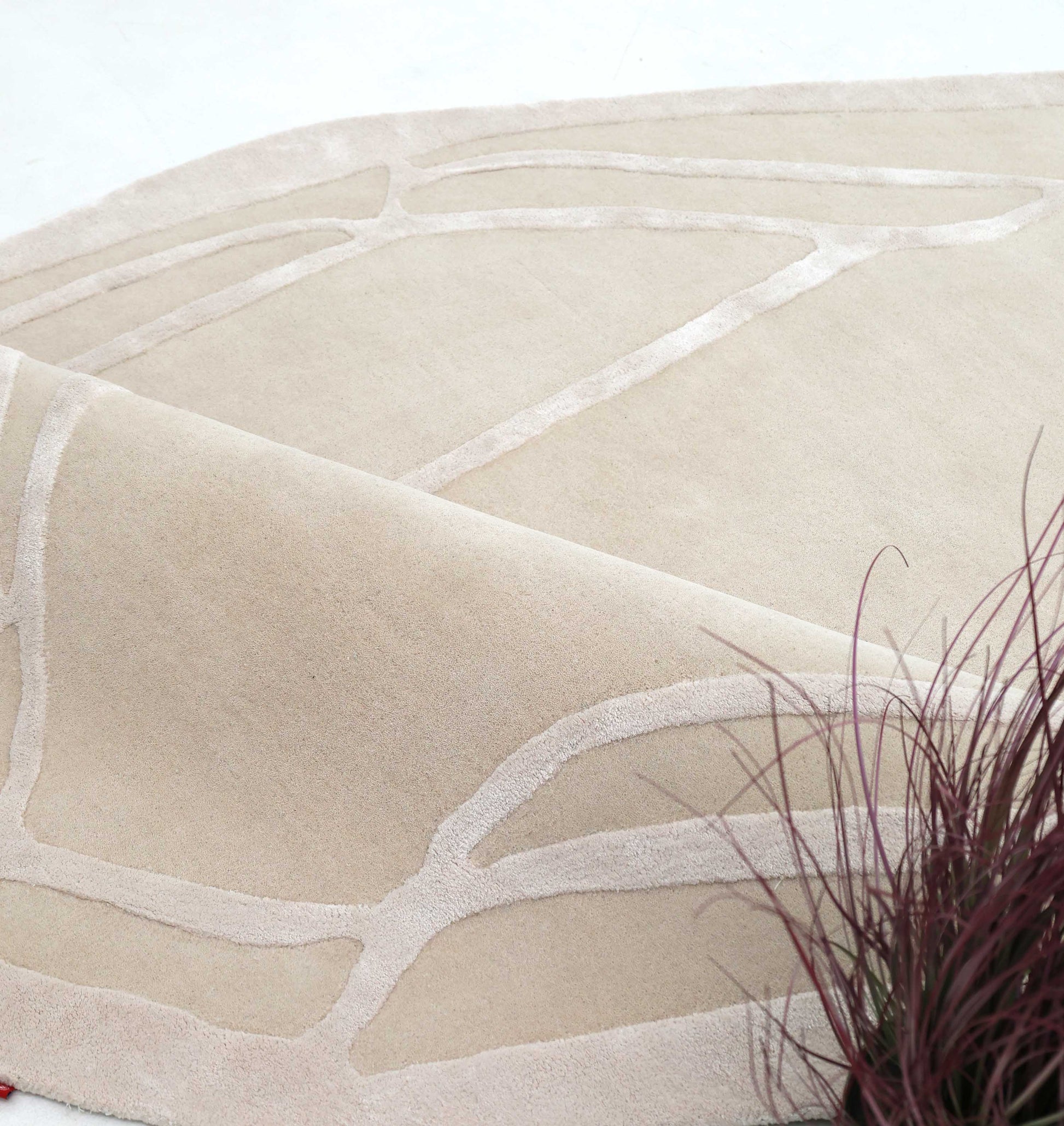 A close-up of the RAMSHA HOME Handmade irregular 200x300 cm Rug HTWV-190, featuring cream and beige geometric patterns with raised abstract lines and a slight corner fold. Reddish-purple decorative grass appears in the front right.