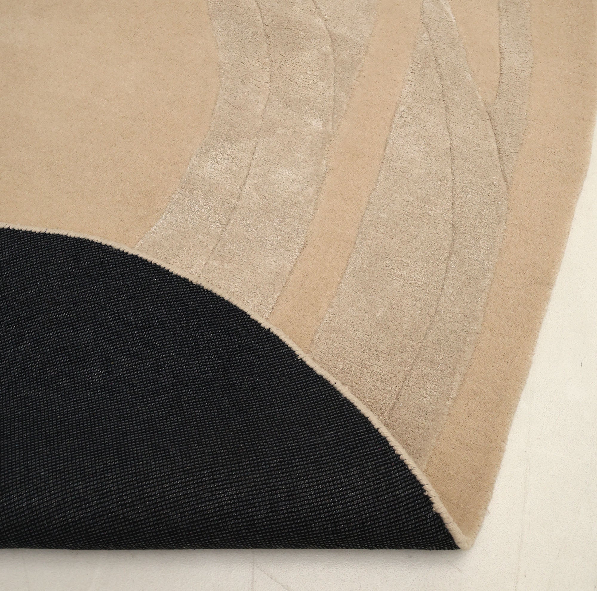 A close-up of the RAMSHA HOME Handmade irregular 200x300 cm Rug HTWV-191 in cream and beige, featuring abstract wave patterns, partially folded to show its black textured underside, displayed on a light-colored floor.