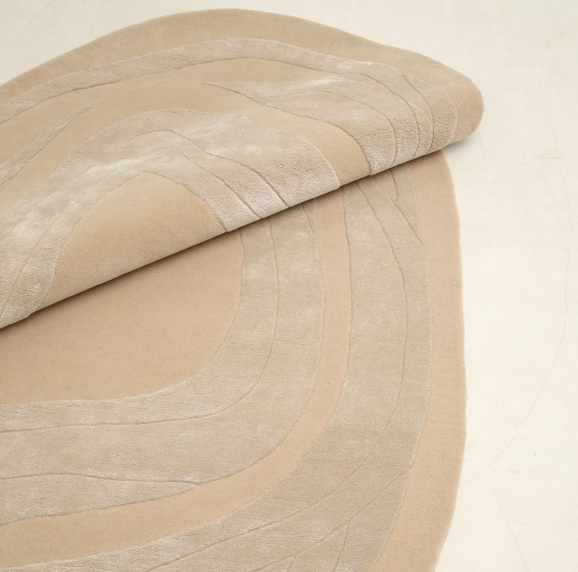 A close-up of the RAMSHA HOME Handmade irregular 200x300 cm Rug HTWV-191, showing its cream and beige tones with raised curving lines. The modern geometric rug is partially rolled to reveal its soft texture on a light surface.