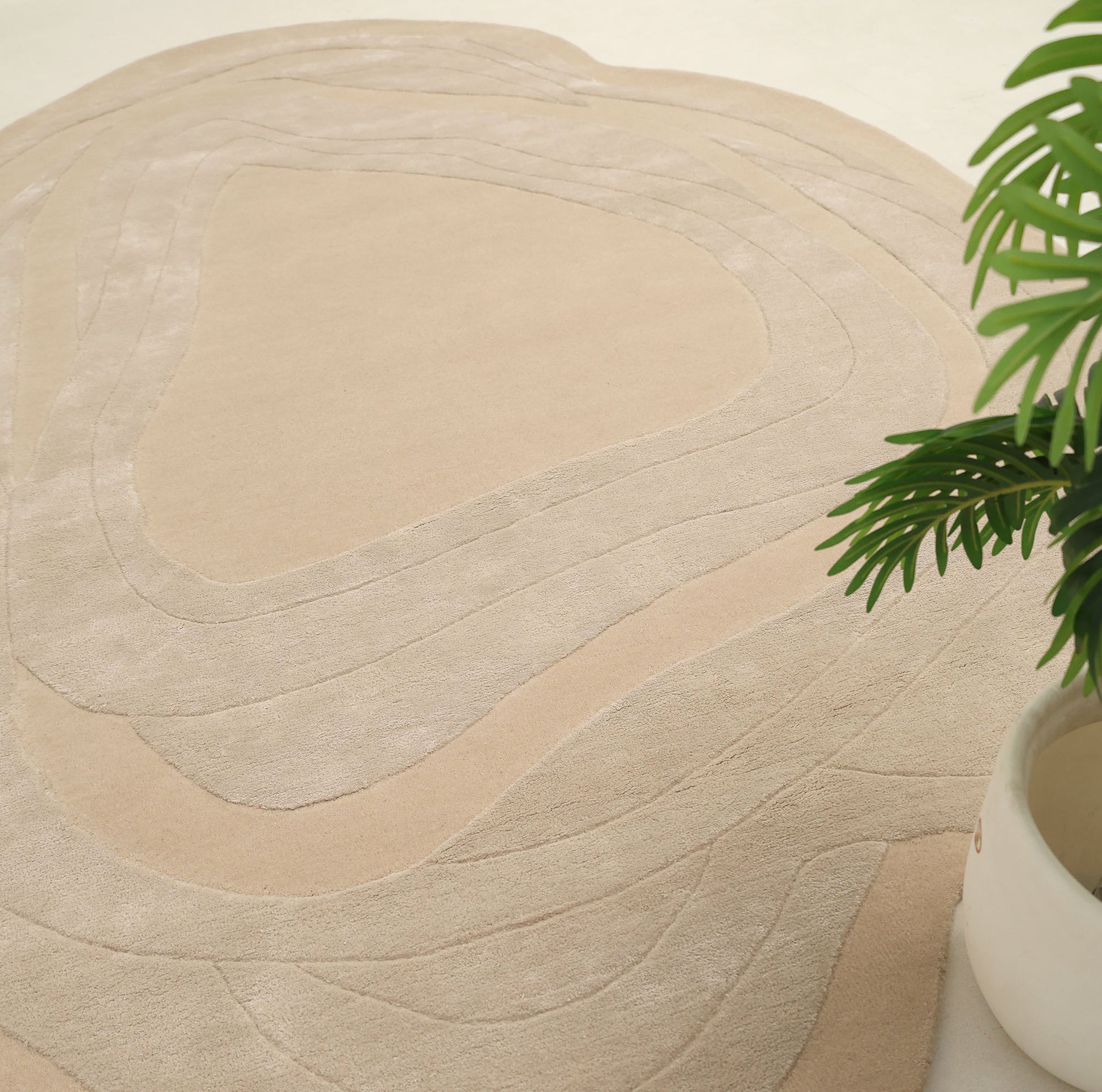 The Handmade irregular 200x300 cm Rug HTWV-191 by RAMSHA HOME features organic, wavy beige patterns. A green plant in a white pot appears partially in the lower right, set on a light neutral floor.