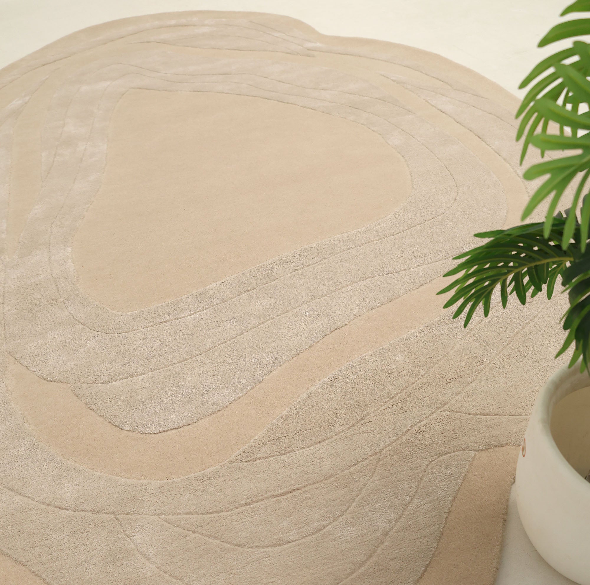 The Handmade irregular 200x300 cm Rug HTWV-191 by RAMSHA HOME features organic, wavy beige patterns. A green plant in a white pot appears partially in the lower right, set on a light neutral floor.