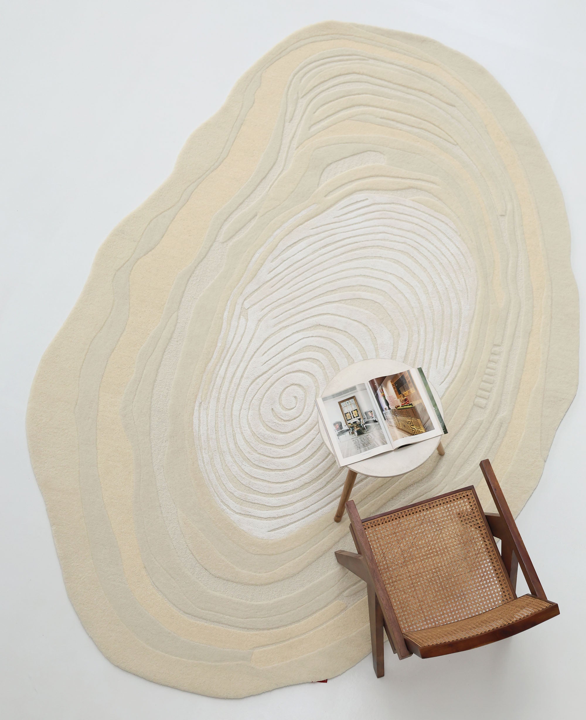A top-down view shows the RAMSHA HOME Ivory Topographic Shaped Rug 200x300 cm (HTWV-192) with concentric circular patterns, a wooden chair with a woven seat, and an open magazine on the uniquely designed rug.
