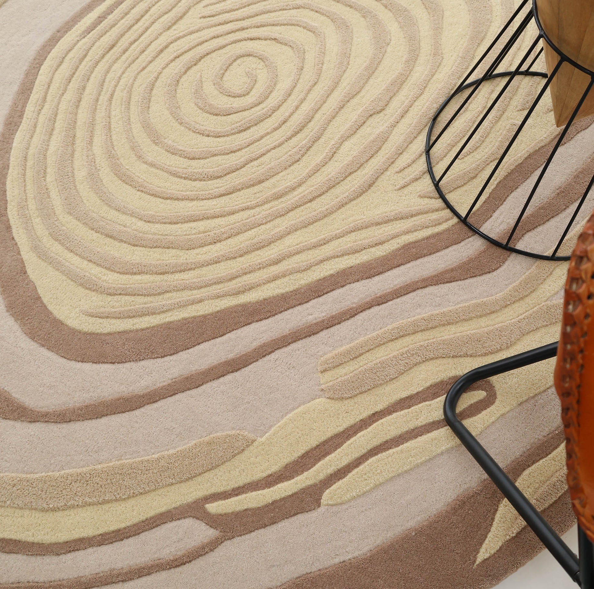 Close-up of RAMSHA HOME’s Terra Eclipse Oval Handmade Accent Rug (HTWV-155) featuring beige, tan, and brown wood grain-like swirls beside a black metal furniture leg and a woven leather chair.