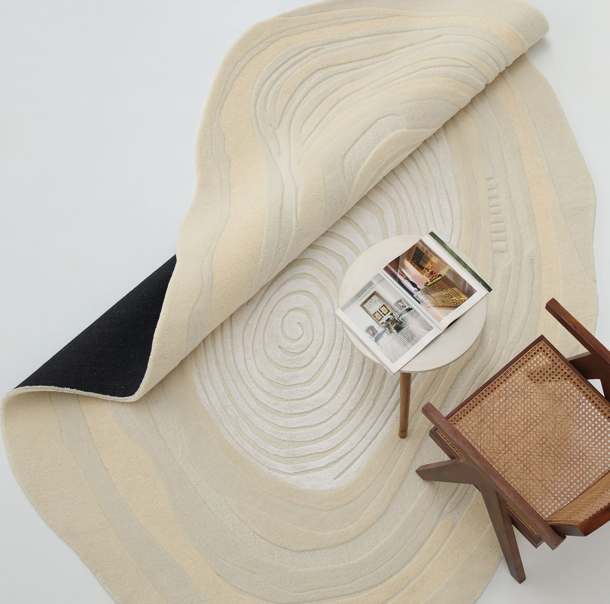 The RAMSHA HOME Ivory Topographic Shaped Rug 200x300 cm (HTWV-192) in cream and beige with a spiral design is partly flipped to show a black underside. A modern wooden chair with a woven seat and a round table with an open magazine rest on the rug.