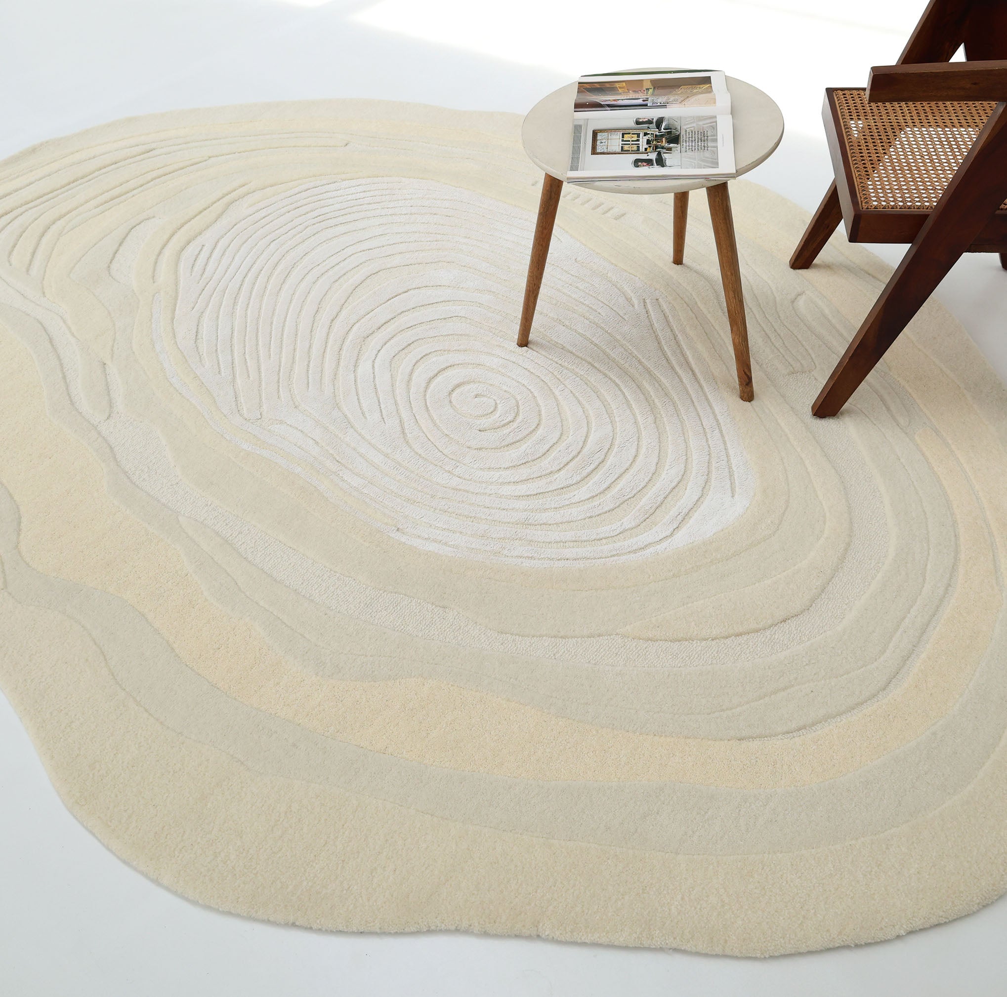 The RAMSHA HOME Ivory Topographic Shaped Rug 200x300 cm (Rug HTWV-192), featuring cream and beige topographic rings, is placed under a round wooden side table with a magazine and a wooden chair.