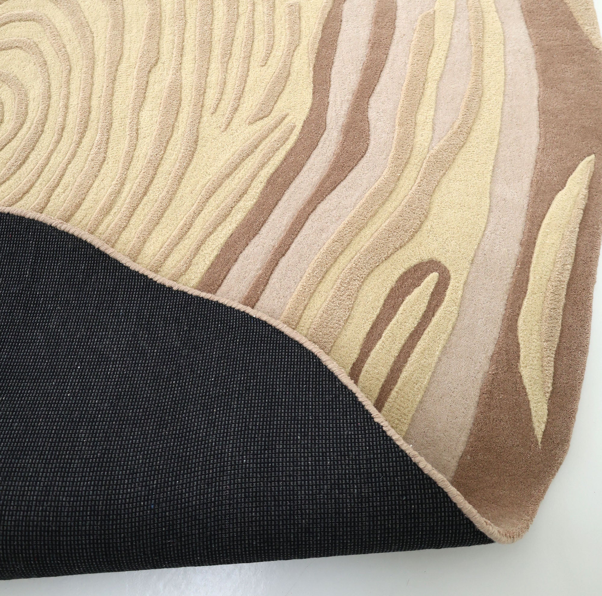 A corner of the RAMSHA HOME Terra Eclipse Oval Handmade Rug Accent Rug HTWV-155 is flipped, showing its black textured underside and wood grain pattern in beige and brown on a white surface.