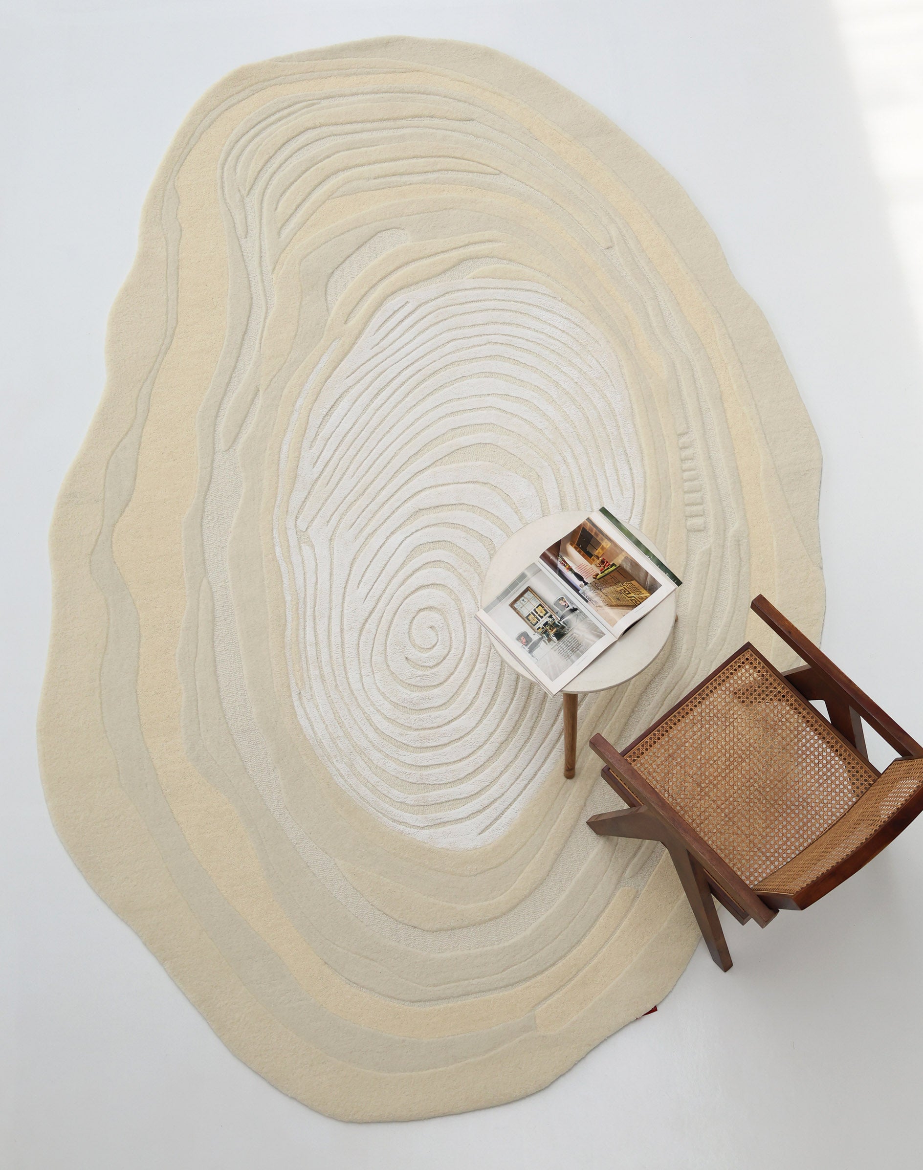 An Ivory Topographic Shaped Rug 200x300 cm by RAMSHA HOME with wavy, wood grain-like lines is paired with a wooden chair with a woven seat and a small white table holding an open magazine.