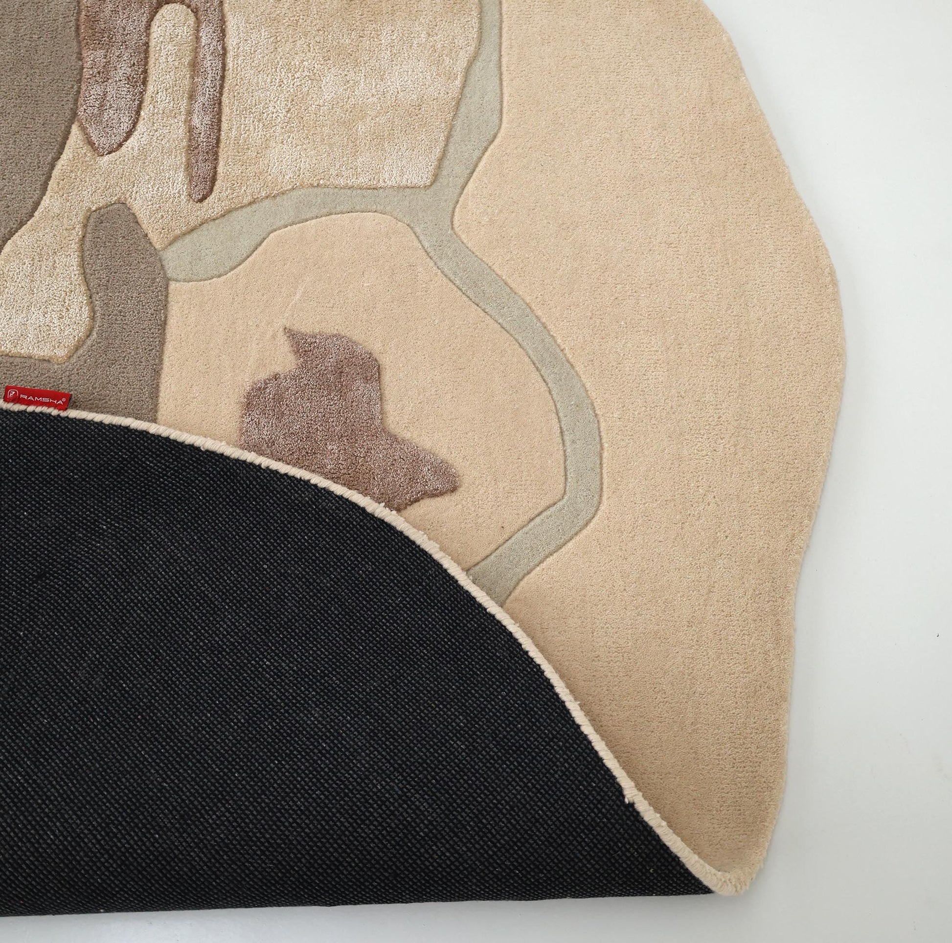 The RAMSHA HOME Round Abstract Shaped Rug with Textured Contour Pattern HTWV-193, in beige and tan, is shown partially flipped on a white floor to reveal its black, textured underside.