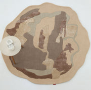 The RAMSHA HOME Round Abstract Shaped Rug with Textured Contour Pattern HTWV-193 features earth-toned beige, brown, and taupe layers with irregular wavy edges. A small round table with a cup and saucer is placed on its left side.