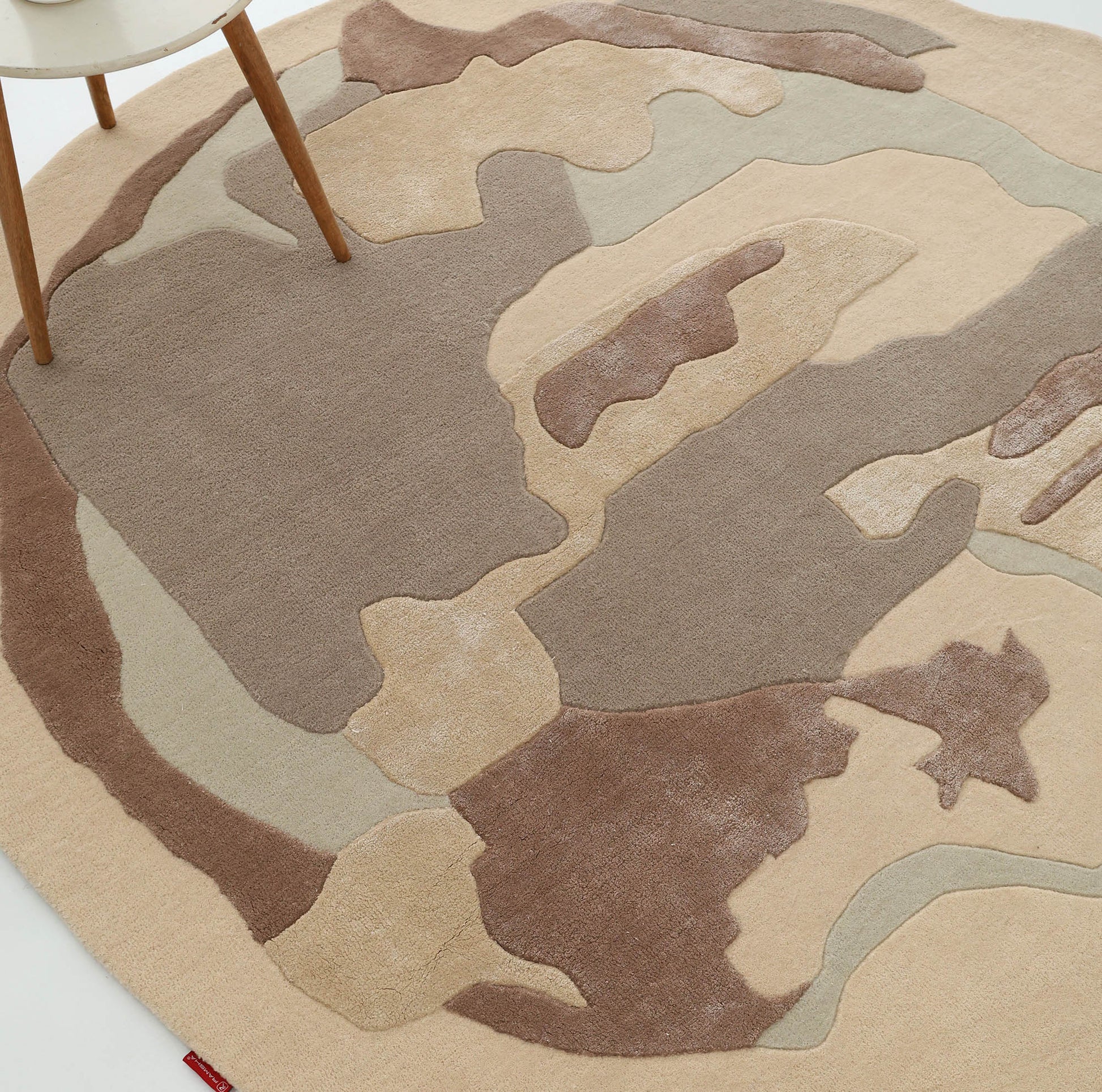 The RAMSHA HOME Round Abstract Shaped Rug with Textured Contour Pattern HTWV-193 in beige, tan, and brown sits on a white floor, with part of a wooden chair visible at the top left.