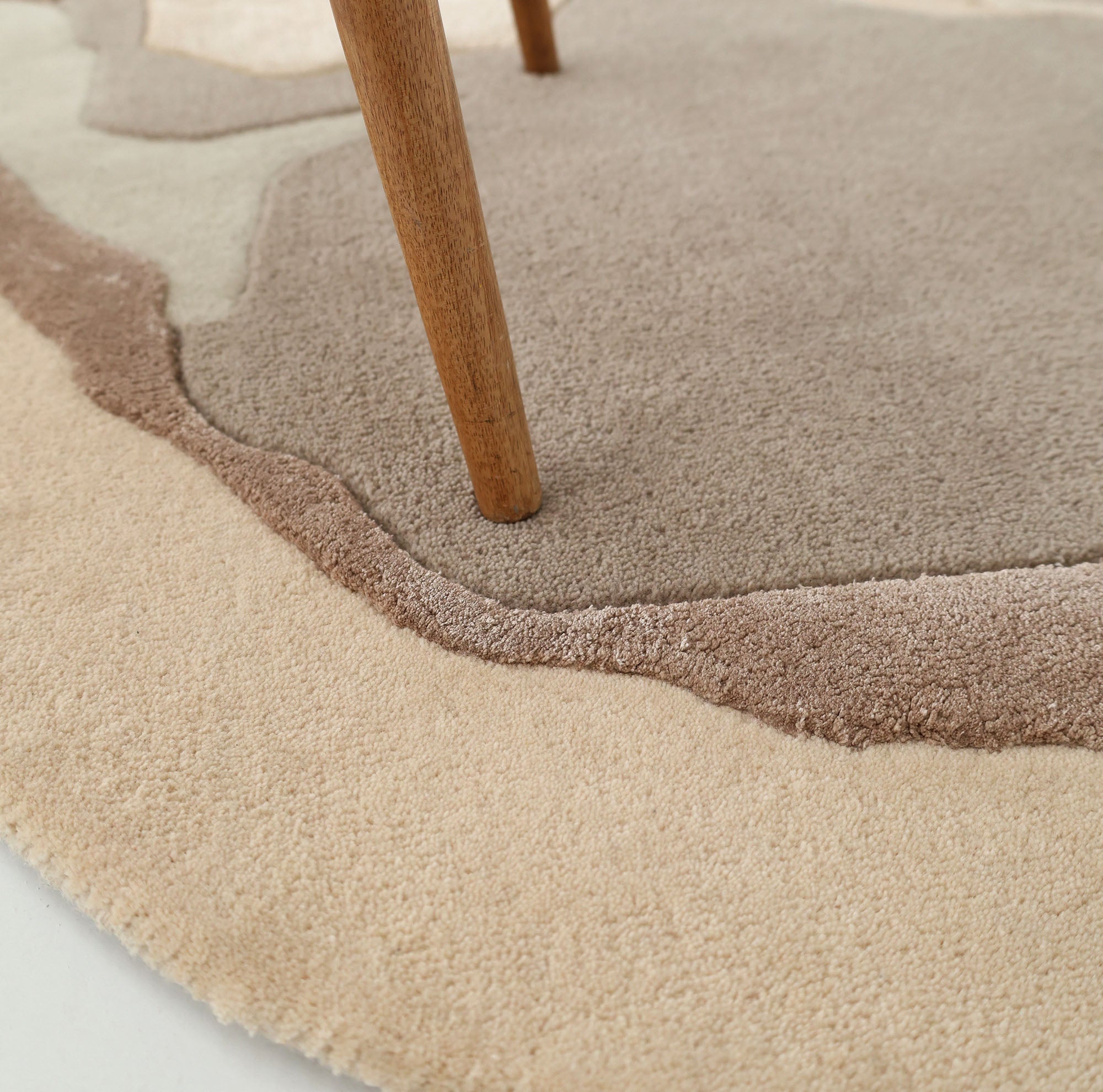A close-up of a wooden furniture leg resting on the RAMSHA HOME Round Abstract Shaped Rug with Textured Contour Pattern HTWV-193, featuring beige, light brown, and taupe wavy designs.