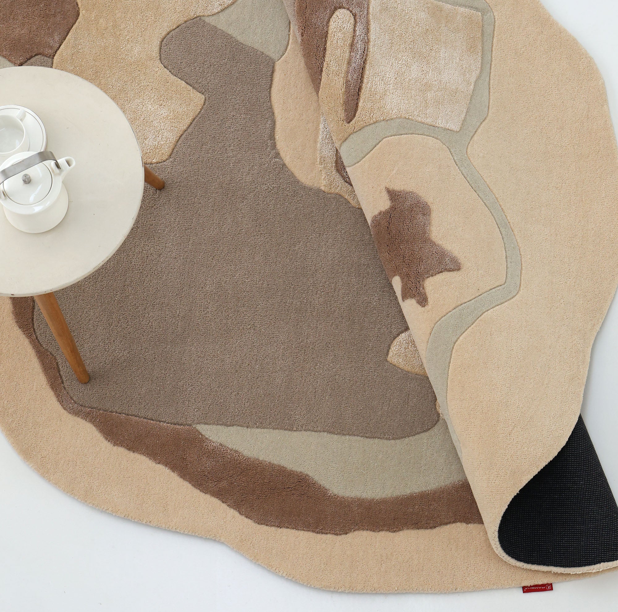 The RAMSHA HOME Round Abstract Shaped Rug with Textured Contour Pattern HTWV-193, in earth tones, is partially folded at the corner. A round, light-colored table with wooden legs sits on the rug, holding a white teapot and cup.