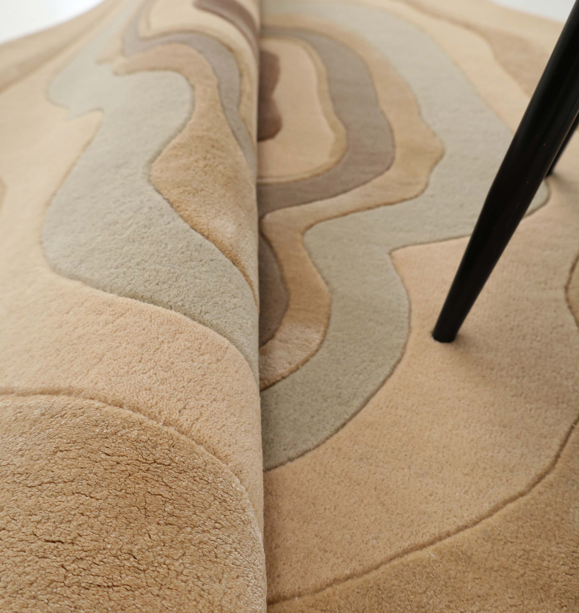 A close-up shows the Luxury Abstract Contour Rug HTWV-194 by RAMSHA HOME, a handmade beige and tan rug with wavy patterns, partially folded up and accented by a black furniture leg resting on it.